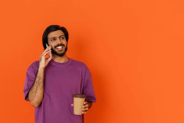 若いですハンサムな笑顔インド人男性でカジュアル紫Tシャツ飲料テイクアウトコーヒー中休憩と電話で話す オレンジの背景に隔離されました コピースペースを見て — ストック写真