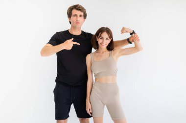 Spor giyimli neşeli, kendine güvenen genç Avrupalı çift, erkek, gri arka planda izole edilmiş kadın pazılarını işaret ediyor. Eğitim sonucu, sağlık, yaşam tarzı, spor ve antrenörle fitness