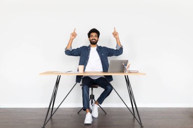 Uzaktan kumanda. Yakışıklı, genç, orta doğulu, günlük kıyafetler giyen, dizüstü bilgisayarda çalışan, serbest çalışan, işyerinde oturan, fotokopi alanını işaret eden ve gülümseyen bir adam.