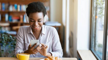 Kulaklıklı neşeli siyahi bayan şehir kafesinde akıllı telefonundan müzik listesi seçiyor. Afro-Amerikalı olumlu bir kadın en sevdiği şarkıyı dinliyor, restoranda, panoramada radyo dinliyor.