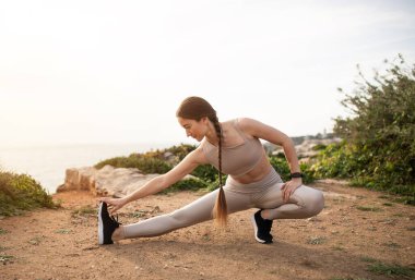 Spor giyimli genç Avrupalı bayan sporcu, deniz sahilinde bacak egzersizi yapıyor. Koşu için ısınma, aktif yaşam tarzı ve motivasyon, açık havada spor