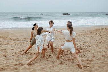 Deniz meltemi ve savaşçı pozları. Spor kıyafetli bir grup genç, okyanus kıyısında yoga yapıyor, daireler çiziyor, sahilde asana egzersizleri yapıyor.