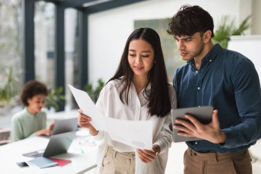 Takım çalışması, İş Arkadaşları Etkileşimi. Modern Ofiste Çalışma Projesini Tartışan Dijital Tablet ve Kağıt Belgeleri Kullanarak Çeşitli İki İş Ekibi. Asyalı kadın ve Arap adam birlikte çalışıyor.
