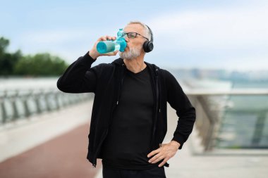 Siyah spor kıyafetleri ve kablosuz kulaklıklar takan motive olmuş yaşlı sporcu dışarıda egzersiz yapıyor. Kıdemli gri saçlı adam briç üzerinde duruyor. Koşarken su içiyor.