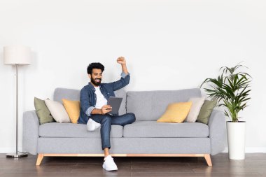 Boş zamanların teknolojisi. Mutlu Arap adam dijital tablet kullanıyor ve evde el kol hareketi yapıyor. Oturma odasındaki kanepede dinlenirken modern aletlerle dinlenen heyecanlı genç bir doğulu. Online kumar