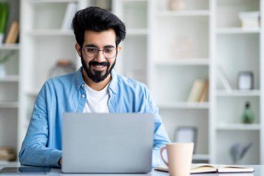 Ofiste dizüstü bilgisayarda çalışan gülümseyen genç Hintli adam, masada oturuyor ve bilgisayar kullanıyor, gözlüklü genç erkek klavyede daktilo ediyor, ekrana bakıyor, uzaktan çalışmanın keyfini çıkarıyor, boşluğu kopyalıyor.
