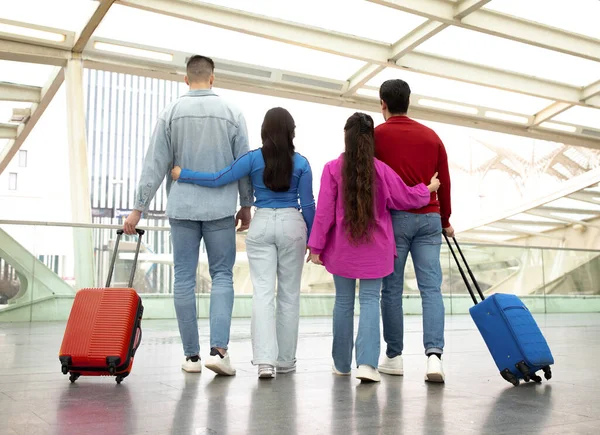 友達と旅行する スーツケースを背負った2組のカップルの裏側には 若い女性が彼氏を抱きかかえ 現代の空港のインテリアで旅行や休暇を楽しんで 一緒に飛行を待っています — ストック写真