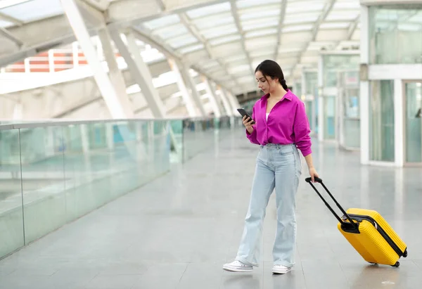 スーツケース付きの携帯電話スタンドを使用してヨーロッパの若い観光客の女性 オンラインテキストと予約チケット モダンな空港の屋内で飛行機を待っている間のウェブサーフィン コピースペース — ストック写真