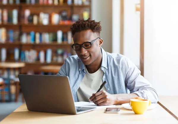 Eラーニングの概念 若いアフリカ系アメリカ人の男は眼鏡を着用し コンピュータのラップトップでカジュアルな服装の学生は カフェで朝食中にノートを取ります ウェビナーに出席 オンラインレッスンを持っています — ストック写真
