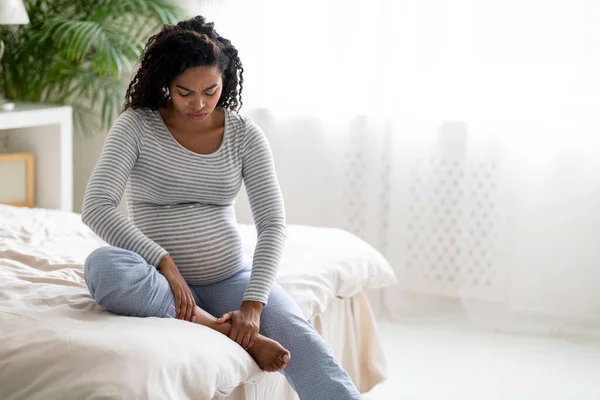 怀孕期间的健康问题 年轻的黑人孕妇穿着家居服 按摩着肿胀的脚 皱着眉头 期待着非洲裔美国女性坐在床上 抄袭空间 — 图库照片