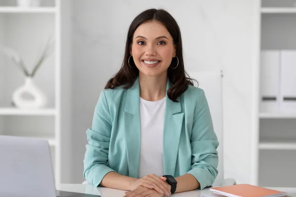Positive confident young european lady manager in suit sit at table workplace in modern office interior. Businesswoman and successful business, video call, work, lifestyle, ad and offer