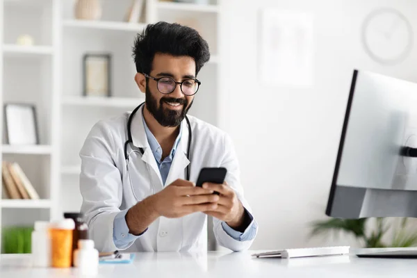 Boldog Fiatal Indiai Orvos Szemüvegben Fehér Kabátban Asztalnál Mobiltelefon Alkalmazásokat — Stock Fotó