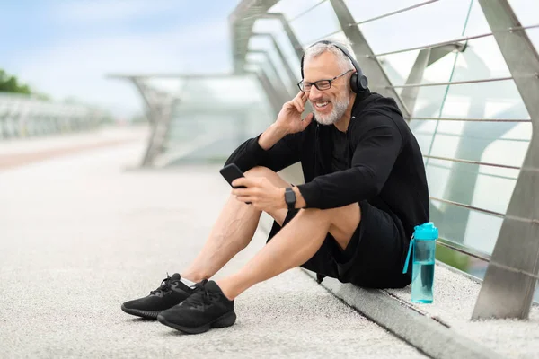 Elderly sportsman sitting on bridge, drinking water, having break while jogging outdoor. Senior athletic man using smartphone, pressing on button on wireless headphones, listening to music