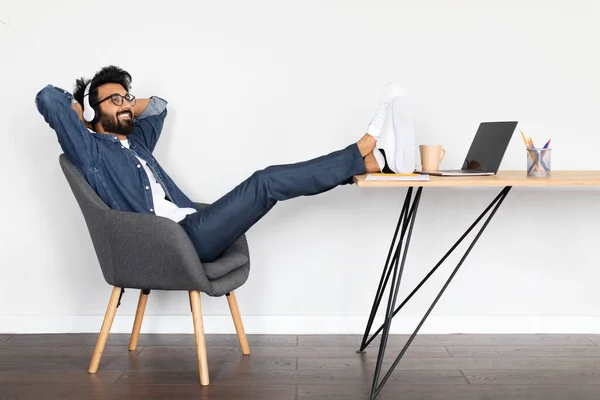 夢のような陽気な若いアラブ人の男のフリーランスは 自宅からオンラインで仕事をしながら休憩し テーブルの上に足でアームチェアでリクライニング ラップトップとワイヤレスヘッドフォンを使用して 音楽を聴く — ストック写真