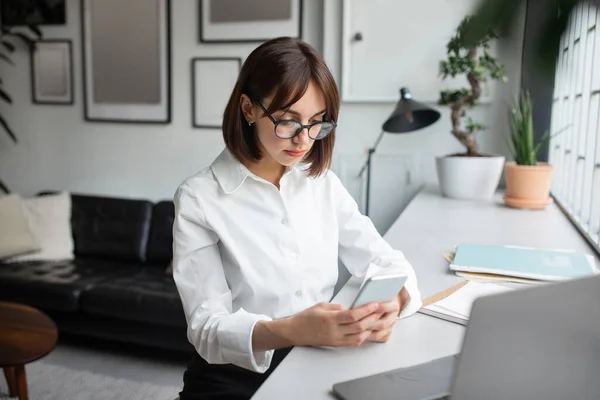 欧洲女性员工在办公室使用手机 女性上网阅读和回复邮件 带着笔记本电脑坐在办公桌前 自由自在 — 图库照片