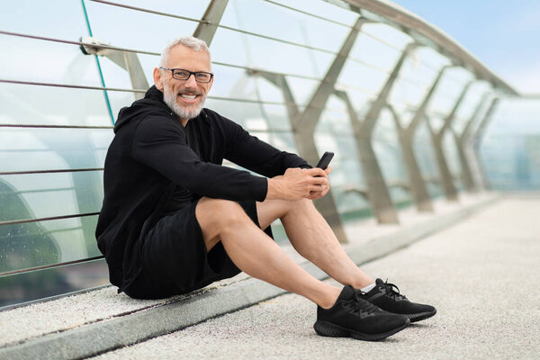 Fitness app. Cheerful mature sportsman handsome grey-haired athletic man in black sportswear having break, sitting on the ground, using smartphone and smiling, jogging by bridge outdoor, copy space