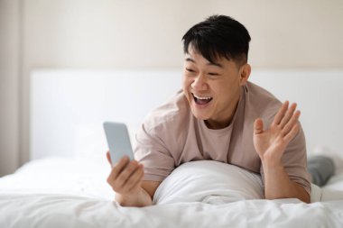 Heyecanlı, orta yaşlı, yakışıklı, pijamalı Asyalı bir adam evde yatar, arkadaşlarıyla video görüşmesi yapar, neşeli bir şekilde gülümser ve akıllı telefon ekranına el sallar, uzayı taklit eder.