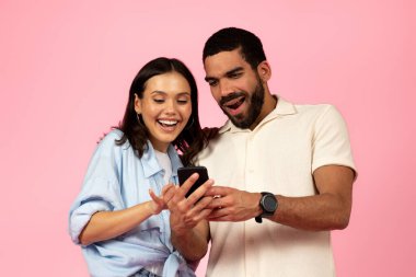 Mutlu, sevgi dolu, çok kültürlü genç çift İspanyol ve esmer kadın pembe renkli stüdyo arka planında akıllı telefon kullanıyor, güzel mobil uygulamaları kontrol ediyor, çevrimiçi asistanı, sohbet robotu...