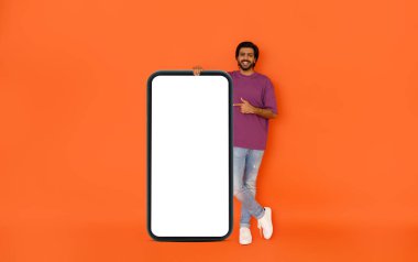 Güzel mobil uygulama. Beyaz ekranlı, dev bir cep telefonunun yanında duran yakışıklı genç adam, başparmağını kaldırıyor, heyecan verici bir online teklif öneriyor, turuncu arka planda izole edilmiş,