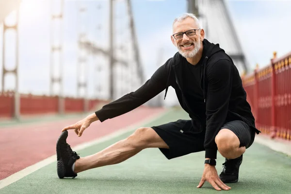 Cheerful healthy elderly man stretching legs and smiling on city bridge. Senior sportsman, outdoor runner, motivation for fitness, energy and active exercise training on the street, copy space