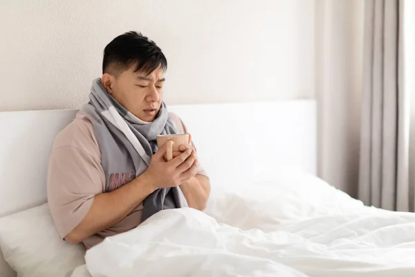 Sick unhappy handsome chinese middle aged man got flu or cold, sitting in bed under blanket at home, wearing warrm scarf around his neck, holding mug, have hot drink, copy space
