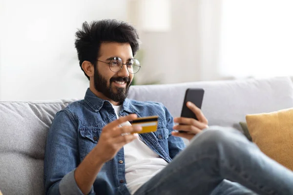 带着智能手机和信用卡在网上购物的英俊的印度人 戴着眼镜的千禧年东方人 喜欢在网上漫游 坐在客厅的沙发上 — 图库照片