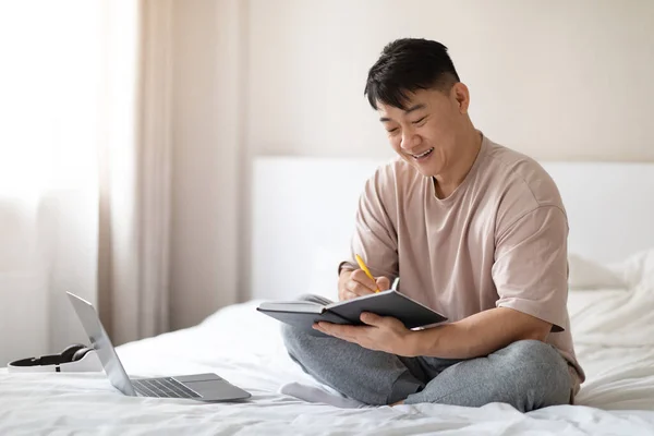 在线教育 网络研讨会 电子学校 穿着睡衣坐在床上 参加培训 使用笔记本电脑 在笔记本上记笔记 复制空间的快乐而又英俊的韩国成熟男人 — 图库照片