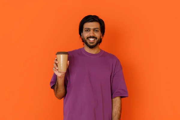 陽気で幸せな若いハンサムな髭を生やしたインド人の男性は 手の中に持っているカジュアルな紫色のTシャツを着て オレンジの背景の上にカメラで微笑んだ ホットドリンク コーヒー — ストック写真