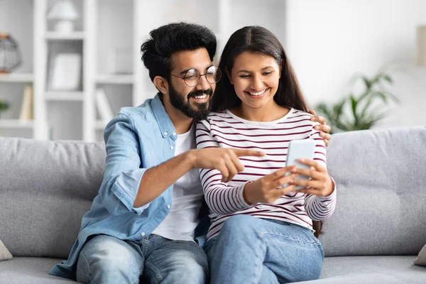 stock image Cute cheerful happy millennial loving indian couple wearing casual outfit sitting on couch in cozy living-room, embracing and smiling, using smartphone at home, shopping online together