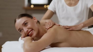 Relaxed young woman enjoying full body massage at new spa salon, professional female masseur massaging her shoulders and back, tracking shot, slow motion, side view