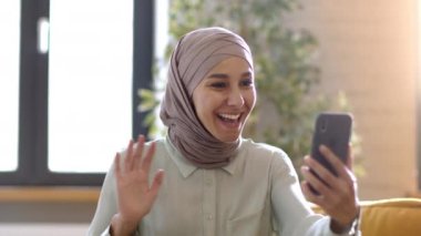 Happy distant communication. Young excited middle eastern lady wearing hijab video calling to her friend via smartphone, waving hand and telling great news. tracking shot, slow motion