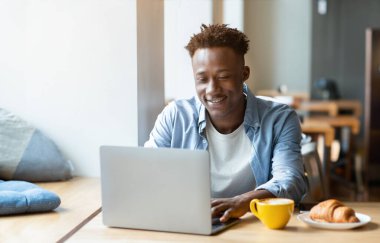 Keyifli, genç siyahi, bağımsız müteahhit şehir kafesinde online iş için dizüstü bilgisayar kullanıyor. Çalışkan genç öğrencinin internet konferansı ya da kahve dükkanında bilgisayar dersi var.
