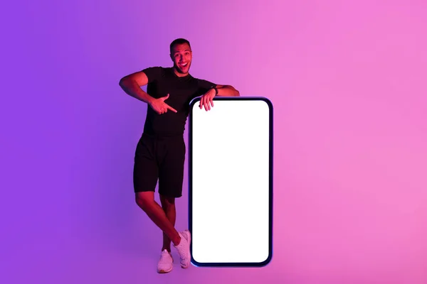 巨大なスマートフォンの近くに立っている若い黒人フィットネス男 空白の画面を持ちます 紫色のスタジオの背景にオンラインワークアウトのためのモバイルアプリケーションを実証デバイスで指を指します モックアップ — ストック写真