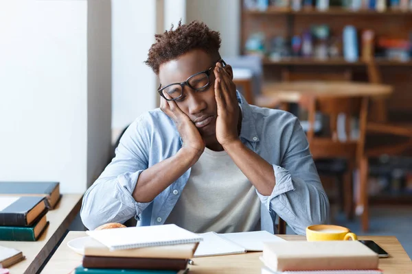 stock image Exhausted tired African American young guy student sitting at desk full of books and notepads, getting ready for complicated exam in university canteen, feeling sleepy, blank space
