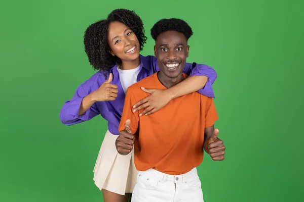 令人高兴的是 千年非洲裔美国女士随意拥抱男友 露出大拇指 享受约会 在绿色工作室背景下与世隔绝 一起批准浪漫和欢乐 人的情感 — 图库照片