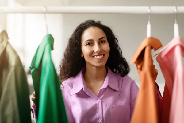 Fashion Business. Cheerful Arabic Woman Shopping Posing With Colorful Clothes On Rack, Smiling At Camera And Choosing New Outfit At Trendy Showroom Store. Choose Your Style Concept