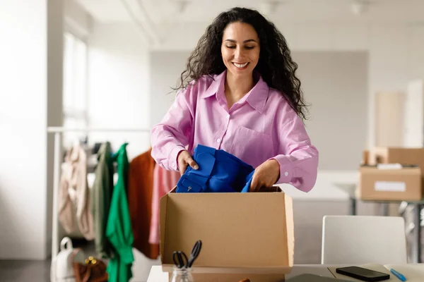 From Fashion Showroom to Shipment. Cheerful Clothing Boutique Owner Lady Packing Trendy Clothes For Delivery, Putting Garment In Cardboard Box At Clothes Showroom Interior