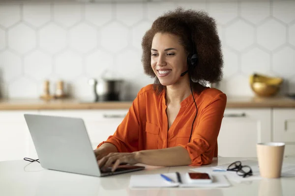 自宅でノートパソコンとヘッドセットでリモートで働く黒人女性を笑顔 キッチンのテーブルに座っている間にコンピュータを使用してアフリカ系アメリカ人フリーランスの女性 仕事を楽しみます — ストック写真