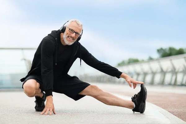 Cheerful healthy elderly man stretching legs and smiling in park with music. Senior sportsman, outdoor runner, motivation for fitness, energy and active exercise training with headhones, copy space