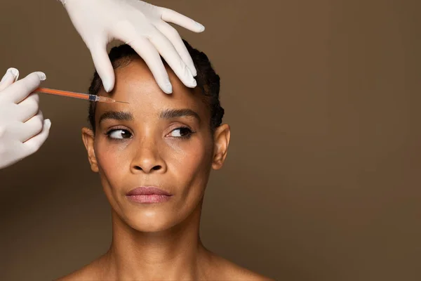美容师Boctor在眉间区注射肉毒杆菌毒素给中年黑人妇女 在棕色工作室背景下接受抗皱治疗 自由自在 — 图库照片