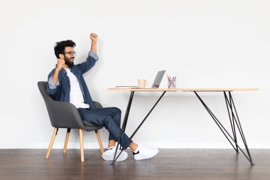 Heyecanlı genç Hintli adam gözlük takıyor dizüstü bilgisayar kullanıyor, el kol hareketleri yapıyor, sohbet ediyor, alışveriş yapıyor, bilgisayar üzerinde çalışan başarılı bir serbest yazar beyaz duvarın üzerinde çalışma masasında oturuyor.