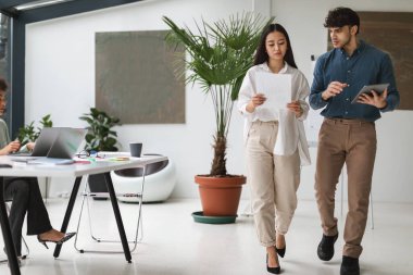 Takım çalışması. Çeşitli iş adamları, Asyalı kadın ve tabletli ve kağıtlı Arap adam modern ofis içinde yürürken konuşup beyin fırtınası yapıyorlar. Tam Uzunluk