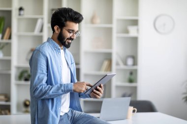İçişleri Bakanlığı 'nda masasının yanında dururken dijital tablet kullanan genç Hintli adam, Gözlüklü Yakışıklı, Gülümseyen Doğu Erkeği Yeni Uygulamaları Gözlüyor, İş İçin Modern Teknolojilerin tadını çıkarıyor, Boşluğu Kopyala