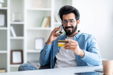 Kolay ödemeler. Gülümseyen Hintli genç adam kredi kartı tutuyor ve içişleri ofisinde otururken cep telefonuyla konuşuyor neşeli doğu erkeği banka müdürüyle konuşuyor, boş yer var.