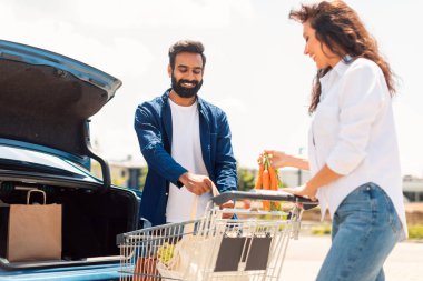 Pozitif ortadoğulu eşler, alışveriş arabaları dolusu ürünlerle birlikte süpermarketin açık hava parkında arabaya doluşuyorlar.