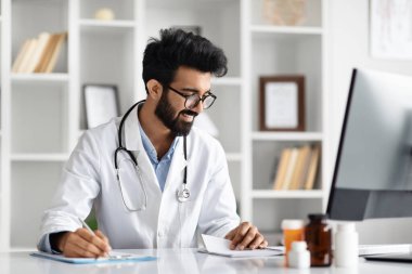 Yakışıklı Hintli doktor bilgisayar kullanıyor ve resepsiyonda oturan tıp dergisine notlar yazıyor. Klinikte çalışan gözlüklü, beyaz önlüklü ve steteskoplu genç doktor.
