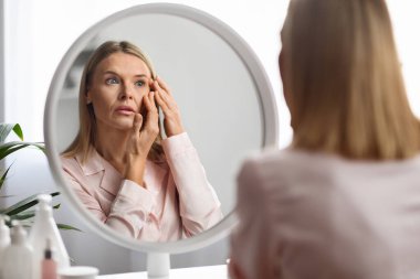 Aging Signs. Upset mature woman looking at wrinkles around her eyes while standing near mirror at home, beautiful middle aged lady inspecting face, feeling stressed, selective focus clipart