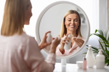 Güzellik rutini. Gülümseyen Orta Yaşlı Kadın Cilt Temizleme İçin Makyaj Sütü ve Pamuk Pedi Kullanıyor, Güzel Olgun Kadın Evde Ayna Duruyor, Günlük Skincare 'ın keyfini çıkarıyor, Seçici Odaklanma