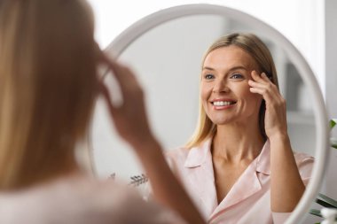 Neşeli olgun kadın evde aynaya bakıyor, göz kremi sürüyor. Göz bölgesi için besleyici serum kullanan çekici orta yaşlı bir kadın. Yatak odasında tuvalet masasında otururken, seçici bir odak noktası.