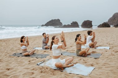 Bir grup mutlu kadın ve erkek spor minderleri üzerinde yoga yapıyor, kumlu sahilde sabahları antrenman yapıyor, bacak bacak bacak üstüne atıp namaste pozu veriyorlar.